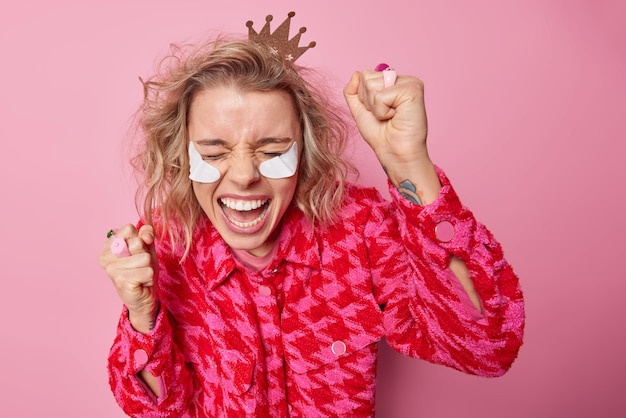 Überglückliche junge Frau bereitet sich auf die Pary vor, trägt Schönheitsflecken auf, tanzt, hält die Fäuste geballt, lacht glücklich, trägt Krone und modische Jacke isoliert über rosa Hintergrund, freut sich etwas