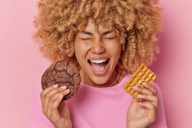 Überglückliche junge Frau mit lockigem Haar hält köstlichen Schokoladenkeks und hausgemachte Waffel hat Naschkatzen hat Zuckersucht, ruft laut aus und hält den Mund isoliert über rosa Hintergrund geöffnet