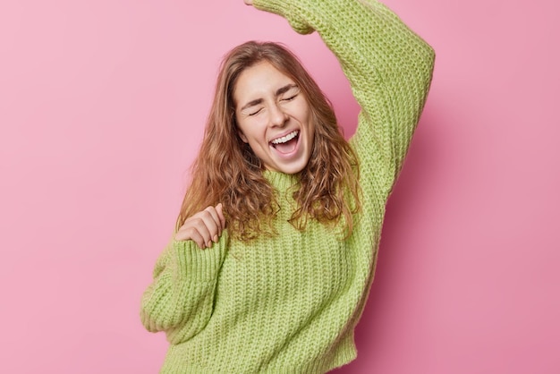 Überglückliche, langhaarige Frau tanzt sorglos, schreit vor Freude, bewegt sich im Rhythmus der Musik, trägt locker gestrickten Pullover, isoliert über rosa Hintergrund, fängt einen schönen Moment ein, hat Spaß und genießt die Freiheit.