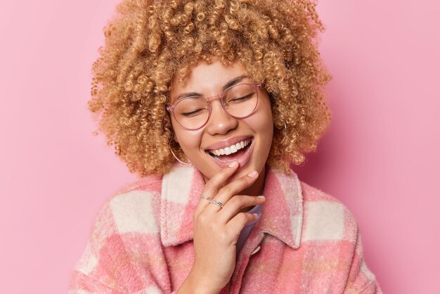 Überglückliche, lockige Frau lacht positiv, lächelt breit und hält die Hand am Kinn. Sie ist gut gelaunt, trägt eine transparente Brille und eine stilvolle Jacke, die über rosa Hintergrund isoliert ist. Fröhliche Emotionen