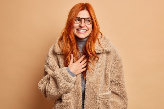 Überglückliche rothaarige erwachsene Frau lacht und drückt glückliche aufrichtige Gefühle aus, trägt eine Brille und einen warmen braunen Pelzmantel, der mit einem Lächeln beiseite konzentriert ist, genießt die Winterzeit und hat eine fröhliche Stimmung. Modekonzept