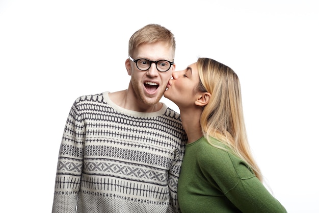 Überglücklicher emotionaler junger bärtiger männlicher Nerd, der Brillen trägt, die aufgeregt ausrufen und schockiert sind, während schöne Frau ihn auf die Wange küsst