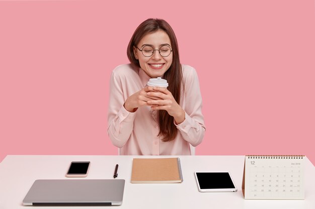 Überglücklicher junger Student hat verträumten Ausdruck, hält Einwegbecher mit Kaffee, hat Pause im Coworking Space, umgeben von modernem Laptop, Tablet