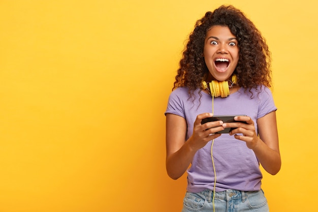 Überglückliches junges Mädchen mit Afro-Frisur, spielt auf dem Smartphone, lacht laut, trägt Stereokopfhörer um den Hals, trägt Freizeitkleidung, isoliert auf Gelb