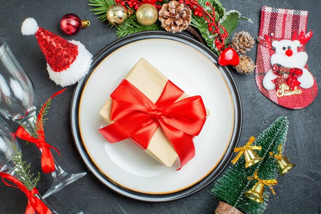 Überkopfansicht der Geschenkbox auf dem Teller der Weihnachtsbaum-Tannenzweige des Nadelbaumkegels Weihnachtsmannhut gefallener Glasbecher auf schwarzem Hintergrund