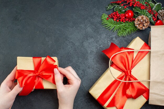 Überkopfansicht der Weihnachtsstimmung mit Hand, die einen der schönen Geschenke und Tannenzweig-Nadelbaumkegel auf dunklem Hintergrund hält