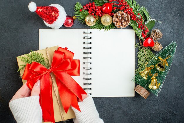 Überkopfansicht der Weihnachtsstimmung mit Tannenzweigen Santa Claus Hut Weihnachtsbaum rotes Band auf Notizbuch auf dunklem Hintergrund
