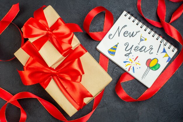 Überkopfansicht von gestapelten schönen Geschenken mit rotem Band und Notizbuch mit Neujahrsschrift und Zeichnungen auf dunklem Hintergrund