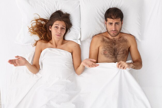 Überkopfaufnahme der verwirrten Frau und ihres Mannes haben sexuelle Probleme im Bett, missfallene Mimik, liegen unter weißer Decke. Der Mensch hat Impotenz, Erektionsstörungen. Tagesleben Familie Probleme Konzept