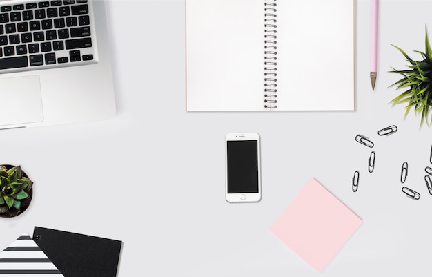 Überkopfaufnahme eines Smartphones auf einem weißen Schreibtisch mit einem Notizbuch, rosa Haftnotizen und Büroklammern