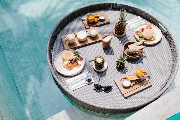 Überkopfaufnahme von Früchten und Süßigkeiten im Pool. Tasse Kaffee und Ananas stehen auf dem Tisch.