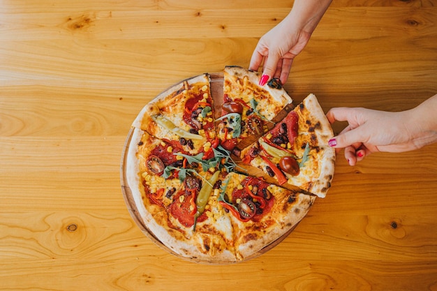 Überkopfaufnahme von zwei weiblichen Händen, die Scheiben von der ganzen Pizza nehmen