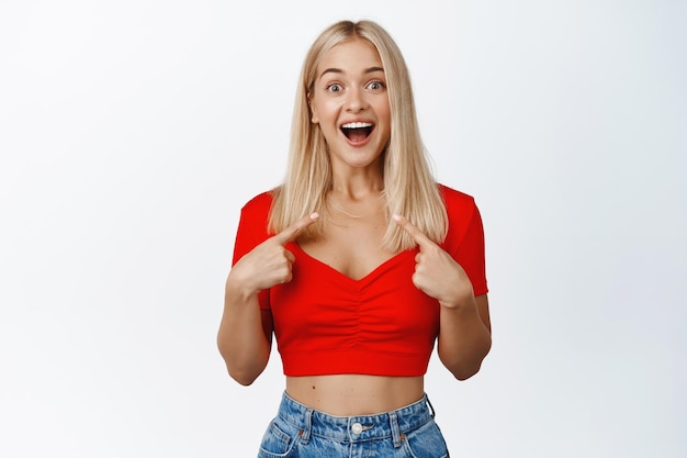 Überraschende blonde Frau, die mit dem Finger auf sich selbst zeigt und aufgeregt lächelt, weil sie ausgewählt wird, erhält großartige Neuigkeiten, die auf weißem Hintergrund stehen