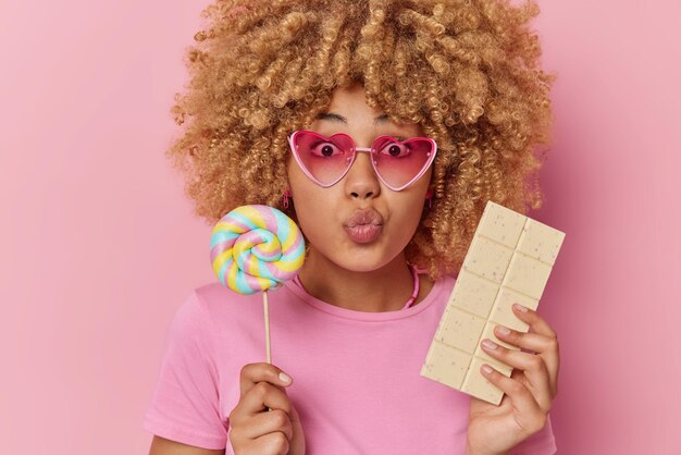 Überraschende junge Frau mit lockigem Haar hält die Lippen abgerundet, trägt eine Sonnenbrille und ein lässiges T-Shirt hält Lutscher und eine Tafel weißer Schokolade isst ungesunde süße Speisen isoliert über rosa Hintergrund