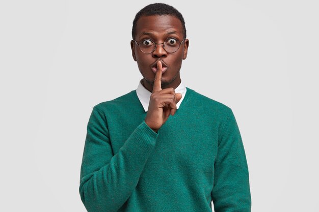 Überrascht attraktiver schwarzer Mann hält Zeigefinger über Mund, zeigt Schweigezeichen, bittet, sein Geheimnis nicht zu verraten