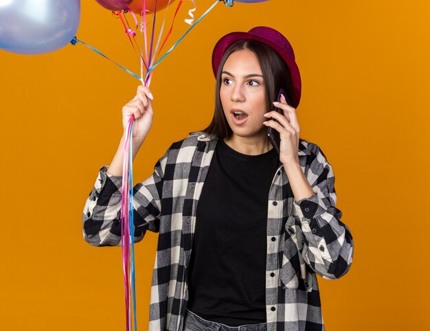 Überrascht auf der Seite junges schönes Mädchen mit Partyhut mit Luftballons spricht am Telefon