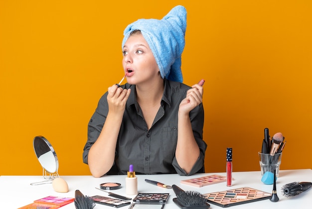 Überrascht aussehende schöne Frau sitzt am Tisch mit Make-up-Tools, die Haare in ein Handtuch wickeln und Lipgloss auftragen