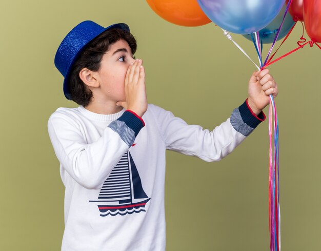 Überrascht aussehender kleiner Junge mit blauem Partyhut, der Luftballons hält, die jemanden anruft