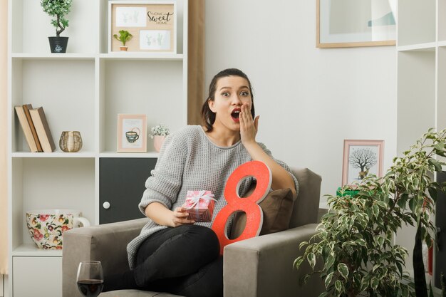 Überrascht bedeckter Mund mit Hand schönes Mädchen am glücklichen Frauentag, der das Geschenk auf dem Sessel im Wohnzimmer hält