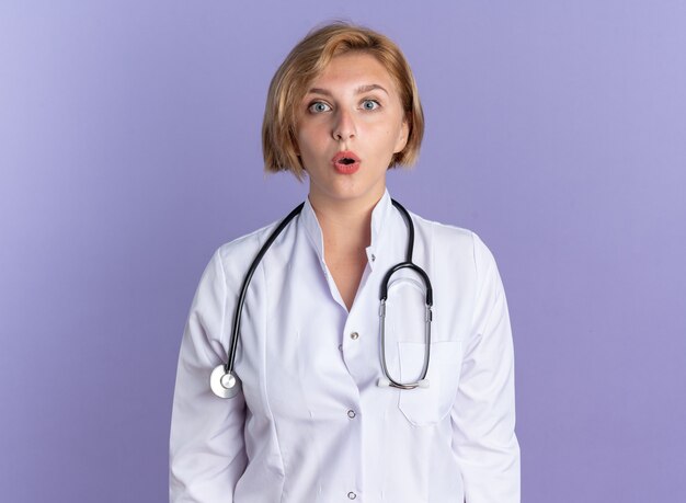 Überrascht Blick in die Kamera junge Ärztin trägt medizinische Robe mit Stethoskop isoliert auf blauem Hintergrund