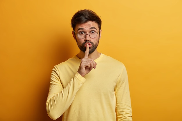 Überrascht drückt der bärtige junge Mann den Zeigefinger an die Lippen, bittet um Ruhe, fordert, kein Geheimnis zu verbreiten, schaut durch eine optische Brille, schaut heimlich, trägt einen gelben Pullover. Shh, sei bitte still