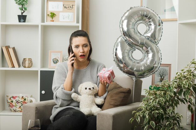 Überrascht, ein schönes Mädchen an einem glücklichen Frauentag mit Geschenk zu suchen, spricht am Telefon, das auf einem Sessel im Wohnzimmer sitzt