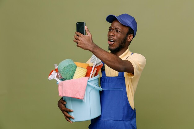 Überrascht, einen Eimer mit Reinigungswerkzeugen zu halten und ein selfie junge afroamerikanische Reinigungskraft in Uniform mit Handschuhen zu nehmen, die auf grünem Hintergrund isoliert sind