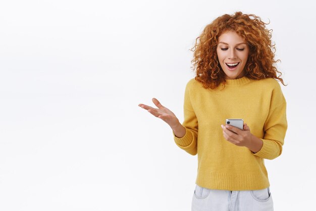 Überrascht, froh, gutaussehend, glücklich, lockiger Ingwer, kaukasische Frau, die eine Smartphone-Lesenachricht vom mobilen Display hält, hebt den Arm, wunderte sich und faszinierte lächelnd, stehender weißer Hintergrund