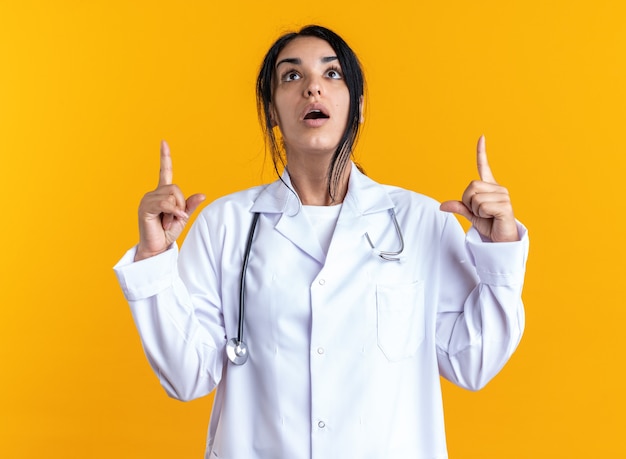 Überrascht, junge Ärztin in medizinischer Robe mit Stethoskoppunkten nach oben isoliert auf gelbem Hintergrund nachschlagen