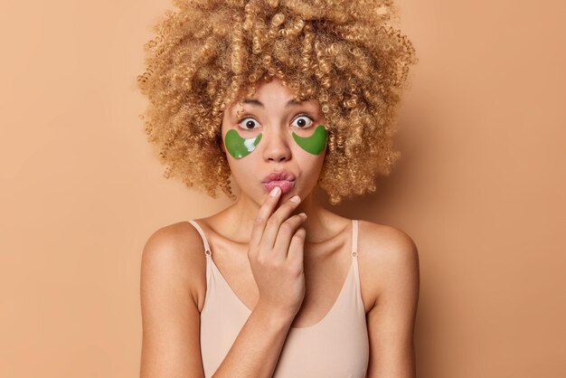 Überrascht, lockige Frau sieht verwundert aus, wenn die Kamera grüne Hydrogel-Patches unter den Augen anwendet, um die Haut zu verjüngen, gekleidet in einem lässigen T-Shirt, das auf beigem Hintergrund isoliert ist. Morgenprozeduren zu Hause