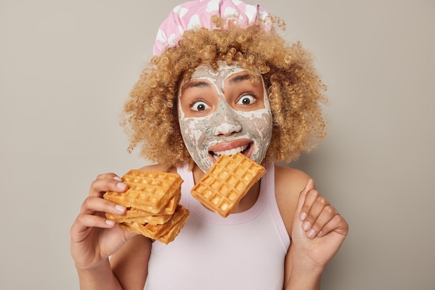 Überrascht lockige Frau trägt Badehut und lässiges T-Shirt beißt Waffel wendet Schönheitsmaske auf dem Gesicht zur Hautbehandlung an, sieht sich isoliert über grauem Hintergrund an Süßes Essen und Wellness-Konzept