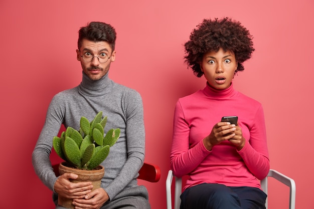 Überrascht schockiert Afro-Amerikanerin benutzt Handy und liest Nachrichten online starrt bugged Augen verwundert Mann posiert in der Nähe auf Stuhl mit Kaktus Topf auf Händen. Interracial Paar innen über rosa Wand