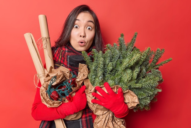 Überrascht schockiert asiatische Frau hält Zweige der Tanne in Papiergirlande für die Dekoration eingewickelt bereitet sich auf das neue Jahr vor trägt Winterhandschuhe kann nicht glauben, dass ihre Augen isoliert über rotem Hintergrund