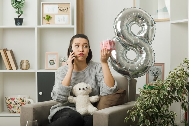 Überrascht, schönes Mädchen an einem glücklichen Frauentag mit Geschenk zu suchen, spricht am Telefon, das auf einem Sessel im Wohnzimmer sitzt