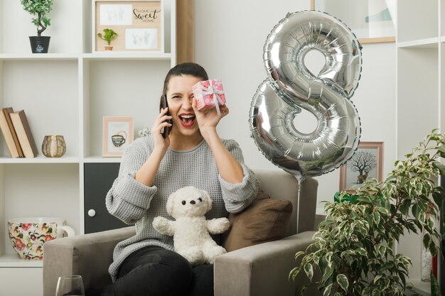 Überrascht, schönes Mädchen an einem glücklichen Frauentag mit Geschenk zu suchen, spricht am Telefon, das auf einem Sessel im Wohnzimmer sitzt