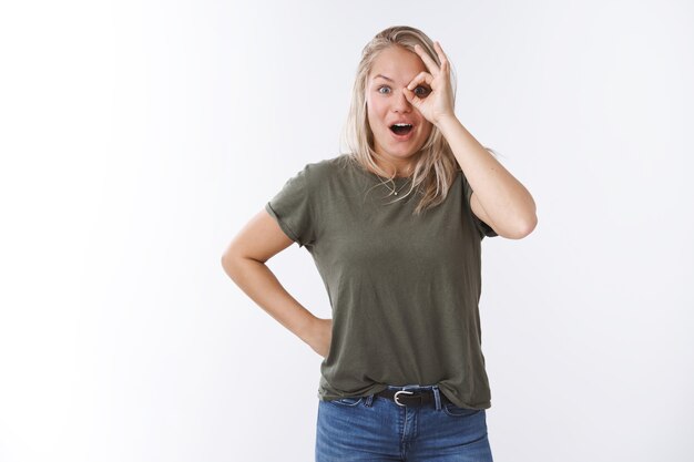 Überrascht und amüsiert, schöne kaukasische Blondine im T-Shirt, die ein okayzeichen über dem Auge zeigt, das den Kiefer keucht, erstaunt von der erstaunlichen überwältigenden Promotion, die aufgeregt über der weißen Wand posiert