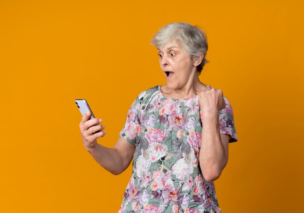 Überraschte ältere Frau, die Faust erhebt, die Telefon lokalisiert auf orange Wand betrachtet