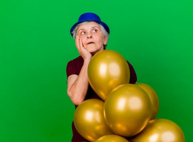 Überraschte ältere Frau, die Parteihutständer mit Heliumballons trägt, setzt Hand auf Gesicht, das Seite lokalisiert auf grüner Wand mit Kopienraum betrachtet