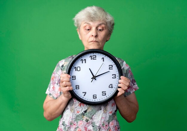 Überraschte ältere Frau hält und schaut auf Uhr isoliert auf grüner Wand