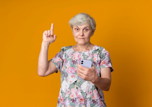 Überraschte ältere Frau zeigt auf, Telefon lokalisiert auf orange Wand zu halten