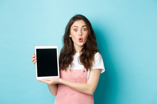 Überraschte attraktive Frau, die einen digitalen Tablet-Bildschirm zeigt und erstaunt in die Kamera schaut, das Promo-Angebot überprüft, amüsiert vor blauem Hintergrund steht