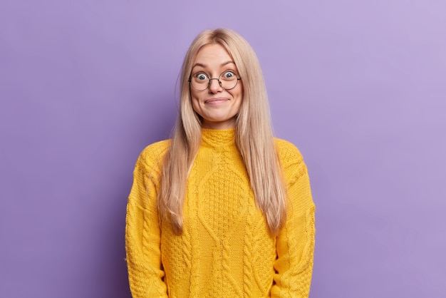 Überraschte blonde europäische Frau hat fröhlichen Ausdruck trägt runde Brille hört unerwartete angenehme Nachrichten in gelbem Pullover gekleidet