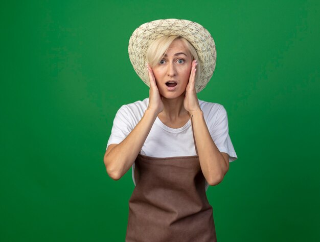Überraschte blonde Gärtnerin mittleren Alters in Uniform mit Hut, die die Hände auf dem Gesicht hält, isoliert auf grüner Wand mit Kopierraum