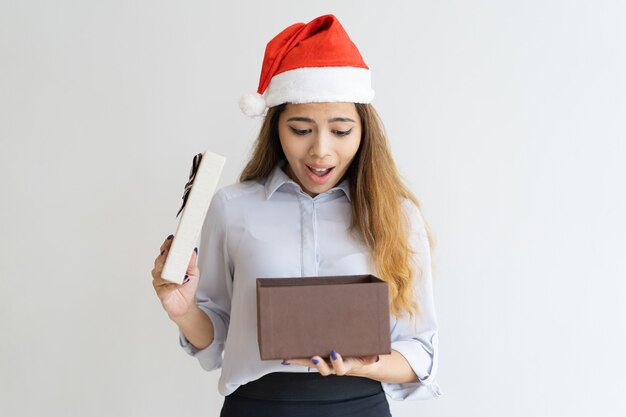Überraschte Dame, die Weihnachtsmann-Hut trägt und in Geschenkbox lugt