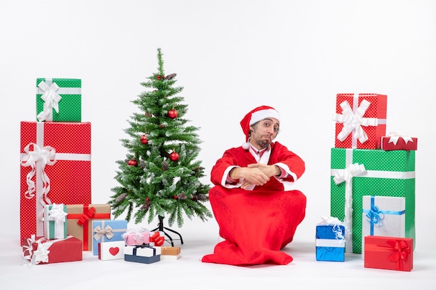 Überraschte emotional aufgeregte junge Erwachsene verkleidet als Weihnachtsmann mit Geschenken und geschmücktem Weihnachtsbaum, der auf dem Boden auf weißem Hintergrund sitzt