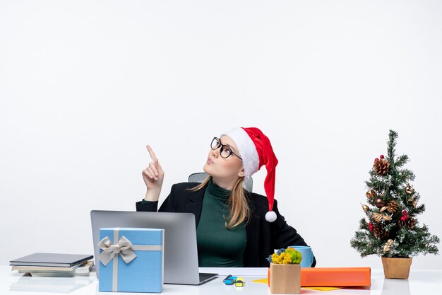 Überraschte ernste Geschäftsfrau mit Weihnachtsmannhut, der an einem Tisch mit einem Weihnachtsbaum und einem Geschenk darauf sitzt, das oben auf weißem Hintergrund zeigt