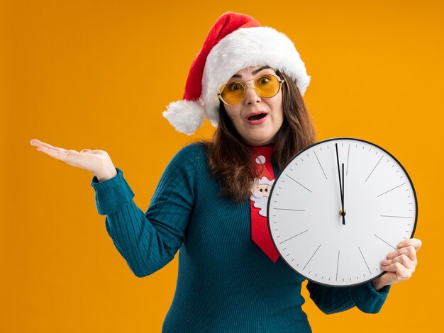 Überraschte erwachsene kaukasische Frau in Sonnenbrille mit Weihnachtsmütze und Weihnachtskrawatte, die Uhr hält und Hand offen hält, lokalisiert auf orange Hintergrund mit Kopienraum