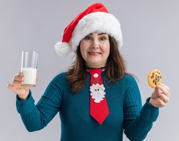 Überraschte erwachsene kaukasische Frau mit Weihnachtsmütze und Weihnachtsmann-Krawatte, die Glas Milch und Keks hält, lokalisiert auf weißem Hintergrund mit Kopienraum