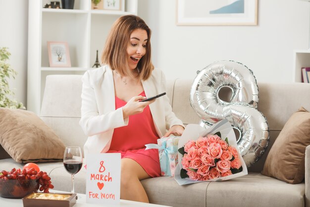 Überraschte Frau am glücklichen Frauentag mit Telefon auf dem Sofa im Wohnzimmer sitzend