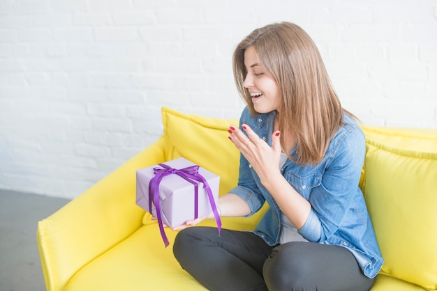 Überraschte Frau, die auf dem Sofa hält Geschenk sitzt